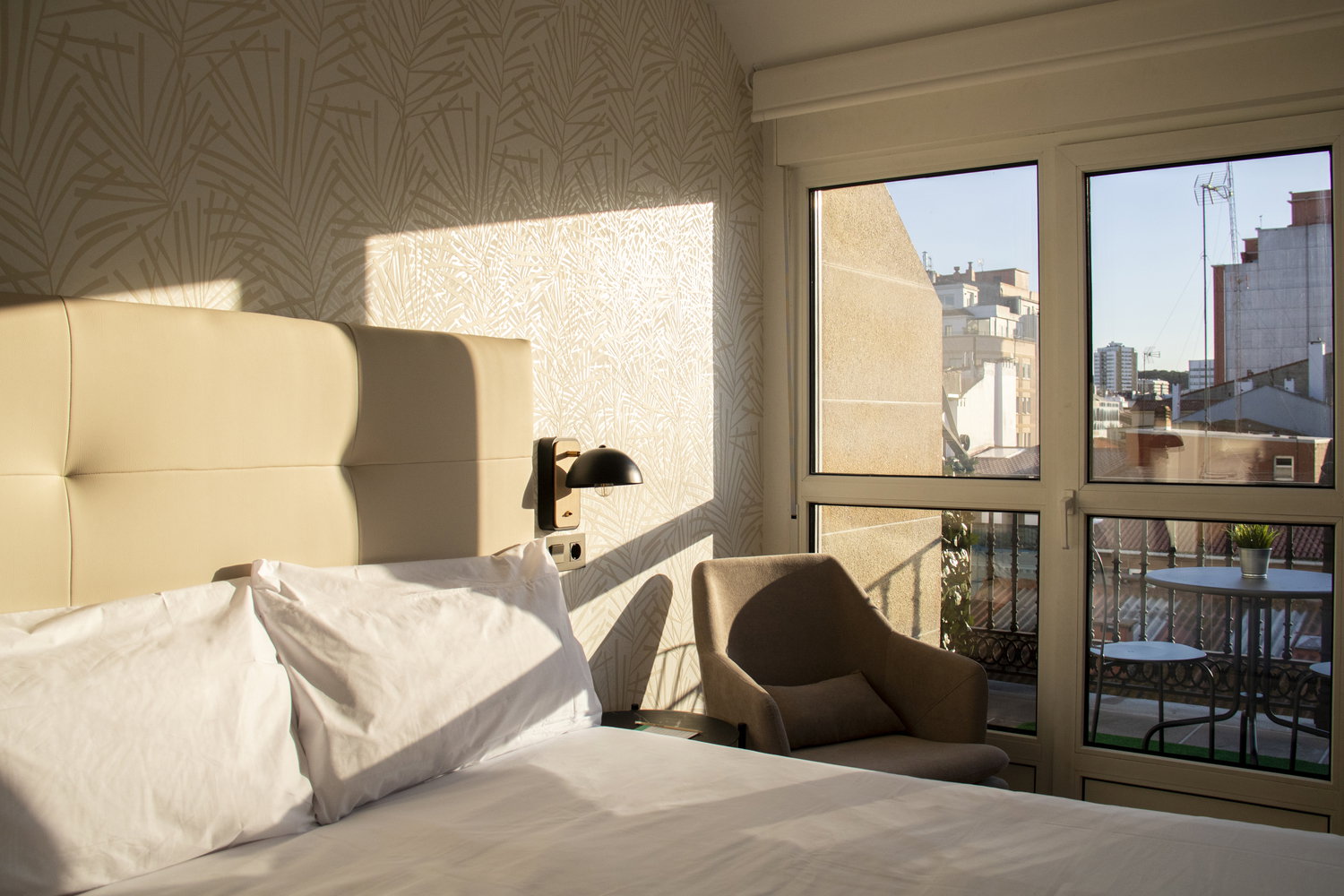 Habitación de hotel iluminada por el atardecer con sillón junto a la ventana y vistas a tejados urbanos