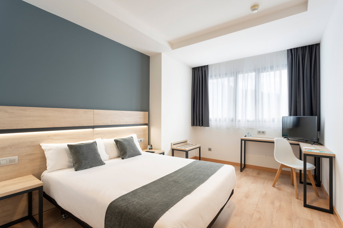 Habitación de hotel con cama de matrimonio, pared azul, escritorio, silla blanca y ventana con cortinas oscuras