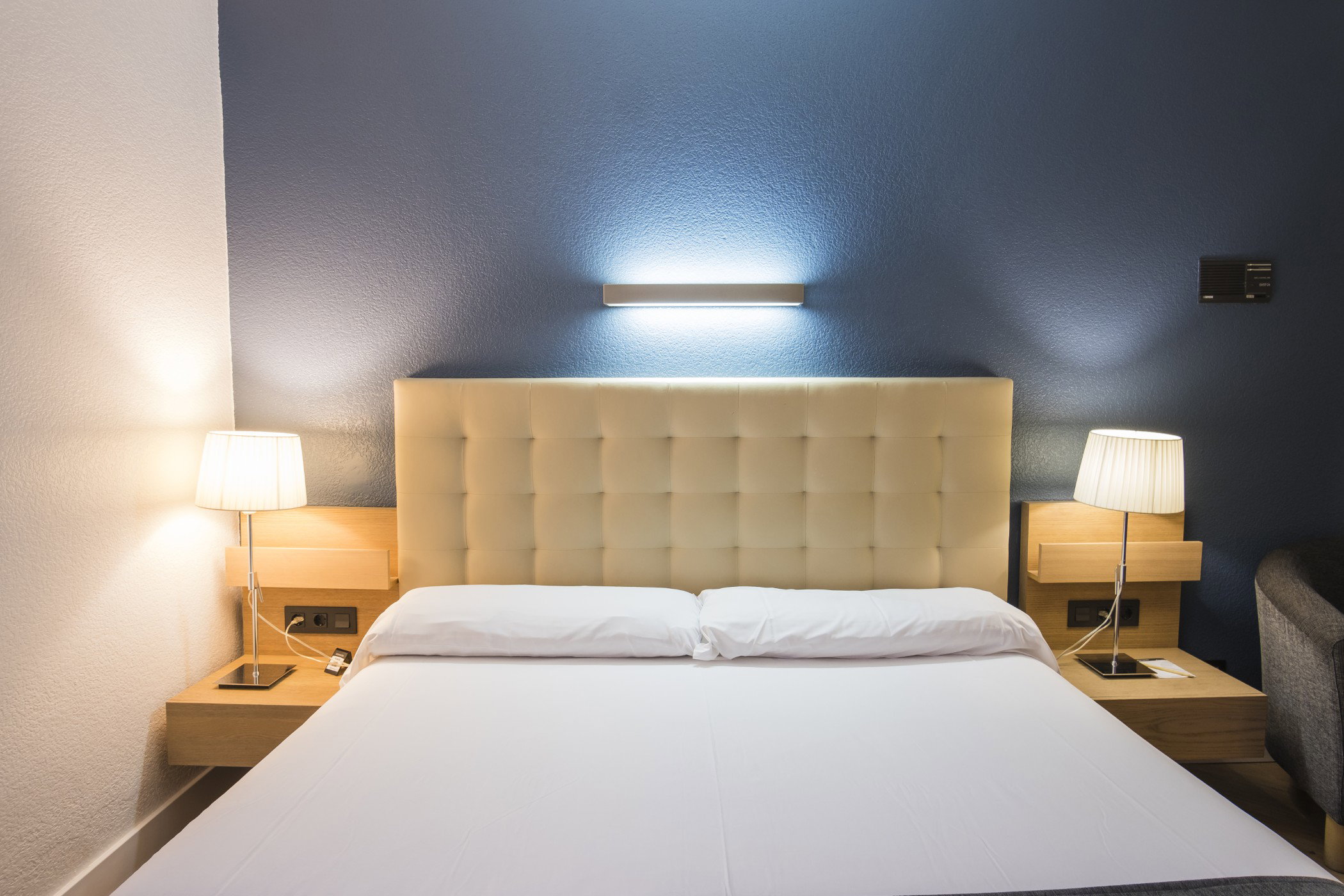 Cabecero acolchado color crema con lámpara de pared encendida sobre una cama con mesillas iluminadas a ambos lados