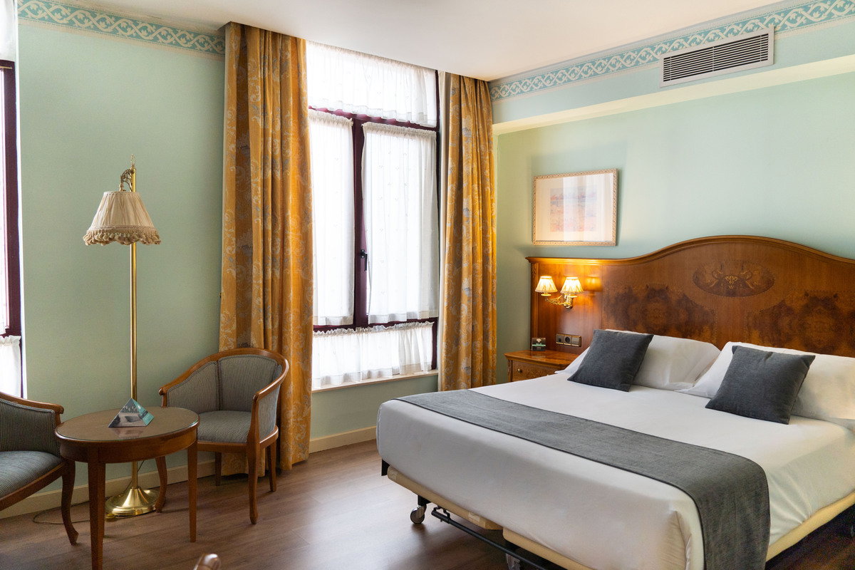 Habitación de hotel de estilo clásico con cabecero de madera, sillones tapizados y cortinas doradas junto a la ventana