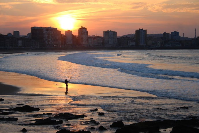 Puesta de sol sobre una playa urbana con la silueta de un pescador y edificios al fondo