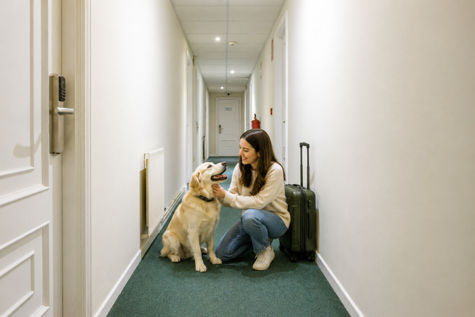 Mujer arrodillada acariciando a un perro golden retriever en un pasillo de hotel, junto a una maleta de viaje