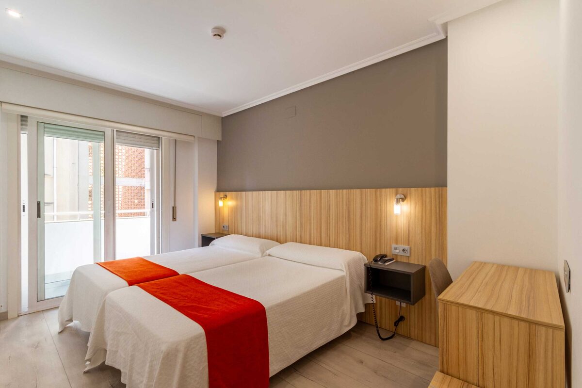 Habitación de hotel con dos camas con cobertor naranja, balcón acristalado y escritorio de madera