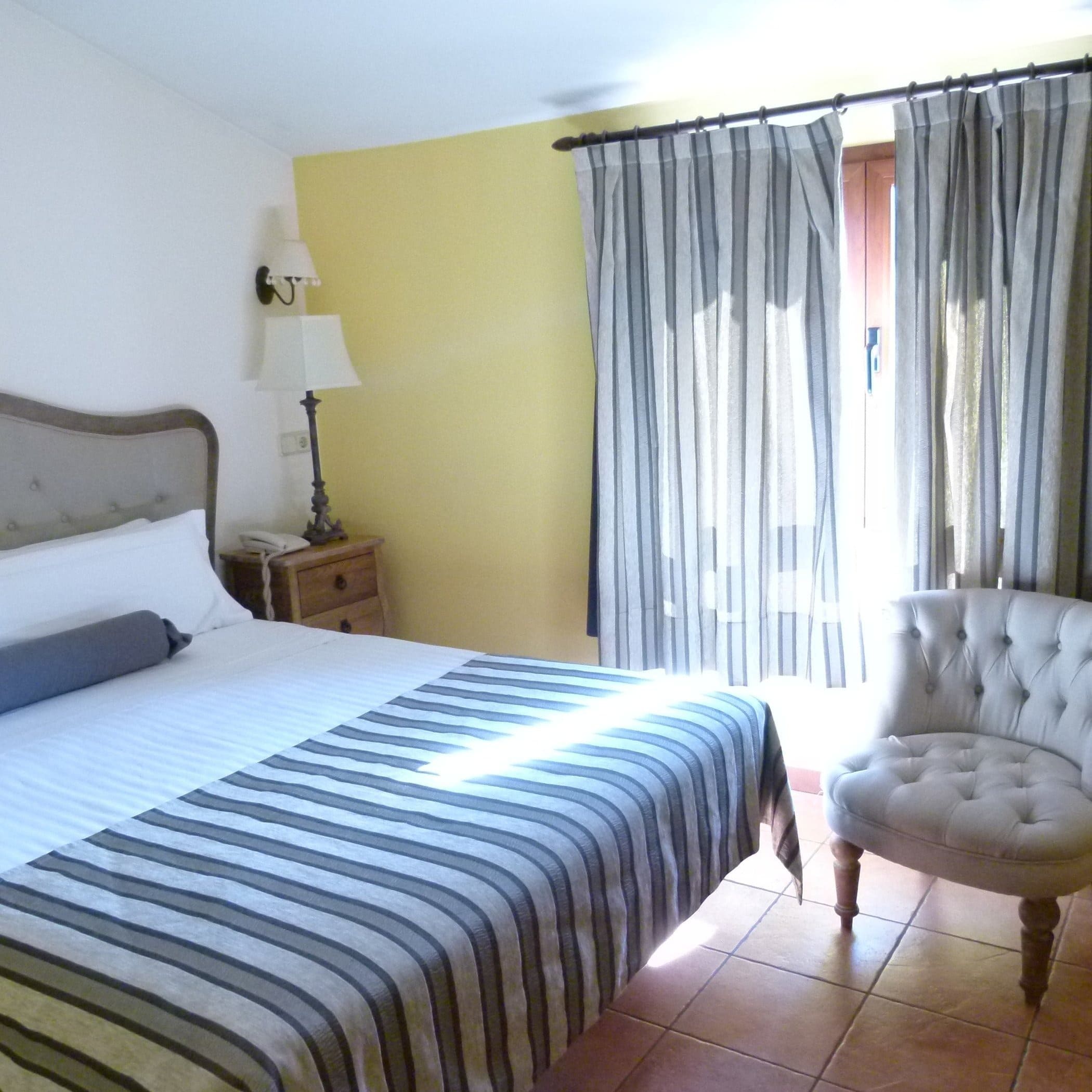 Habitación con cama de rayas azules, pared amarilla, sillón tapizado y ventana con cortinas