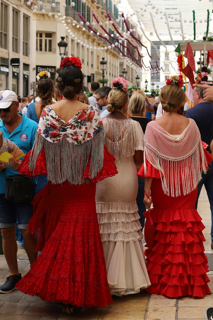 Tres mujeres de espaldas con trajes de flamenca y mantones de flecos caminando por una calle decorada con guirnaldas