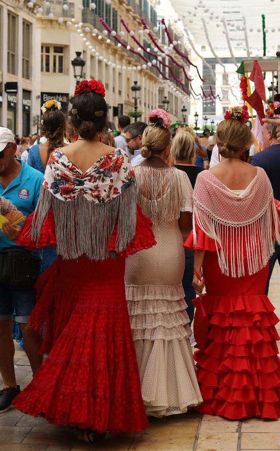 Tres mujeres de espaldas con trajes de flamenca y mantones de flecos caminando por una calle decorada con guirnaldas